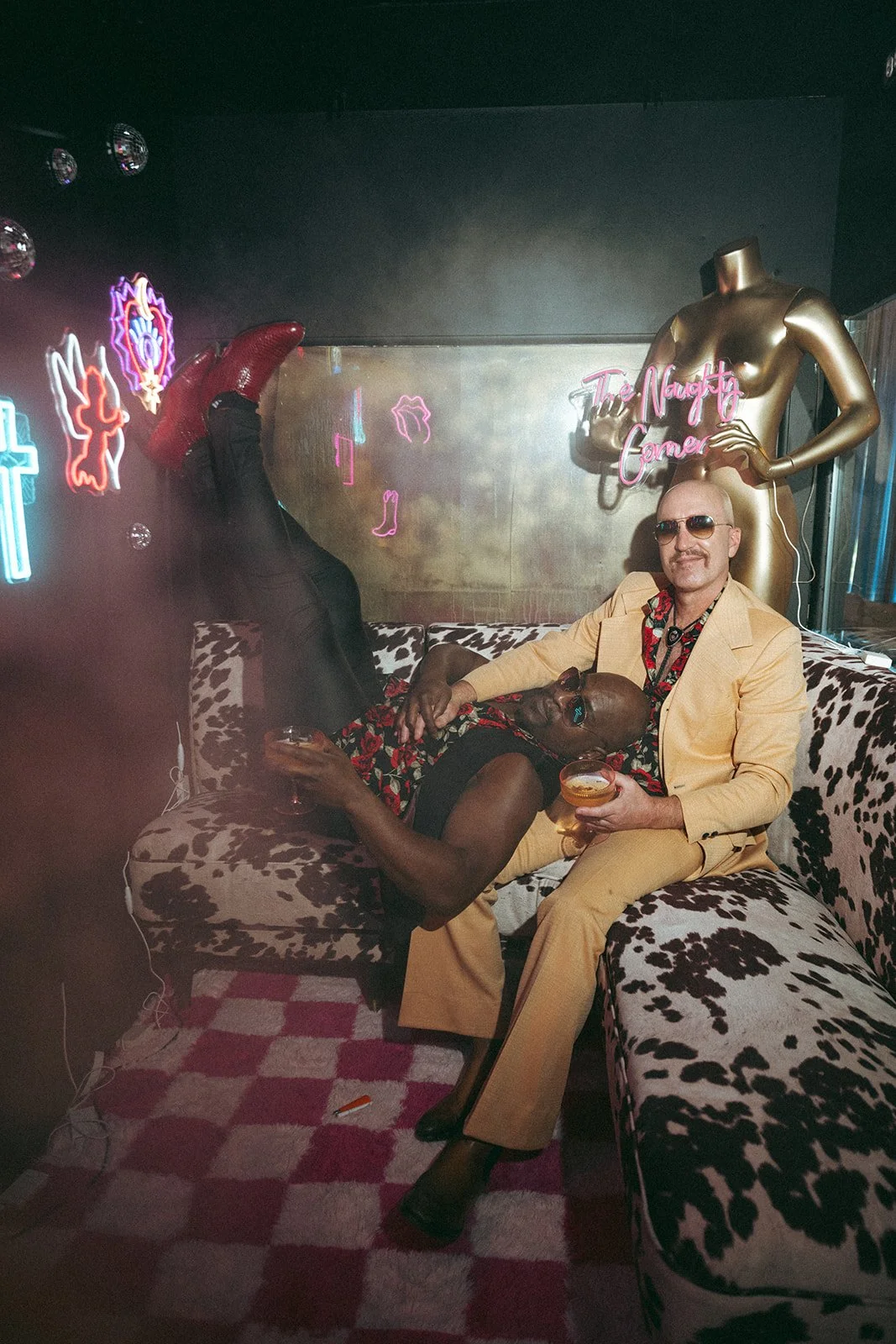 A man in a beige suit and sunglasses sitting on a leopard-print couch, with a person in a floral dress lying across his lap. The person is holding a cocktail and wearing red boots. The background features neon signs and a gold mannequin with a neon sign reading 'The Naughty Corner'.