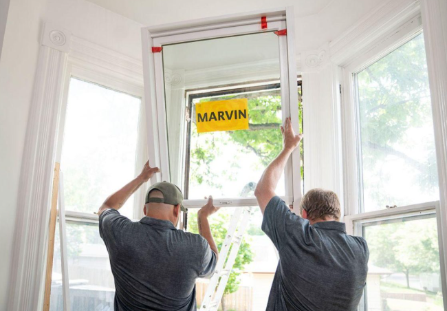 Two men installing a window screen frame inside a house with large windows and a tree outside. One man is wearing a baseball cap, and the other has gray hair. A yellow sign with the name 'MARVIN' is attached to the window.