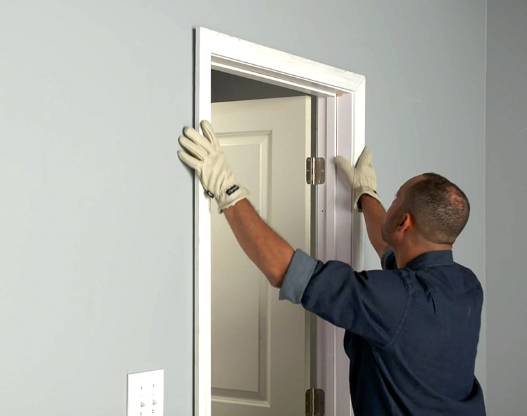 A man installing or adjusting a doorframe without a door, wearing gloves, in a light-colored room.