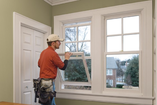 A man installing a window screen in a room with light green walls and large double-hung windows.