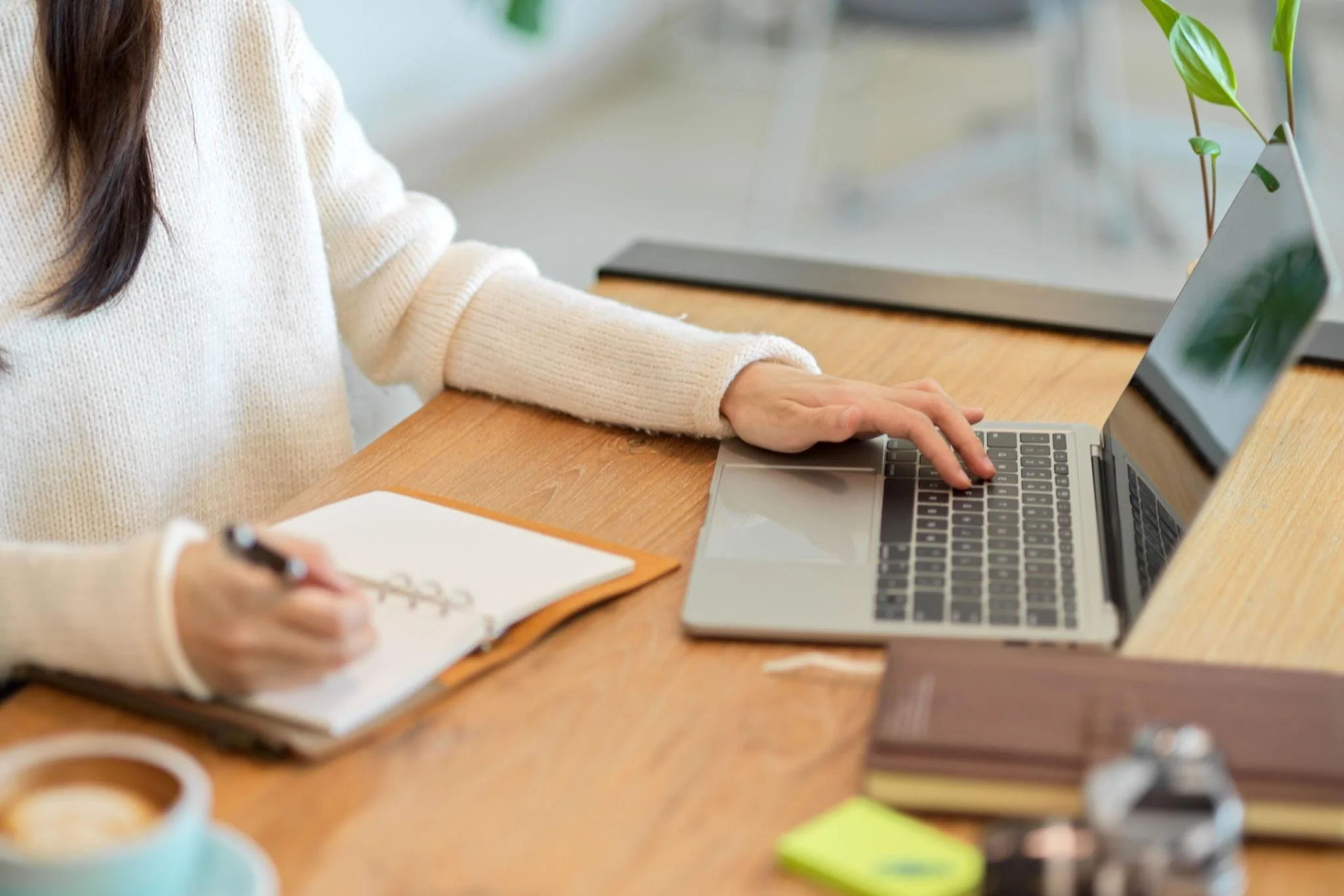Person in a cream-colored sweater using a laptop at a wooden desk, writing in a notebook, with office supplies around.