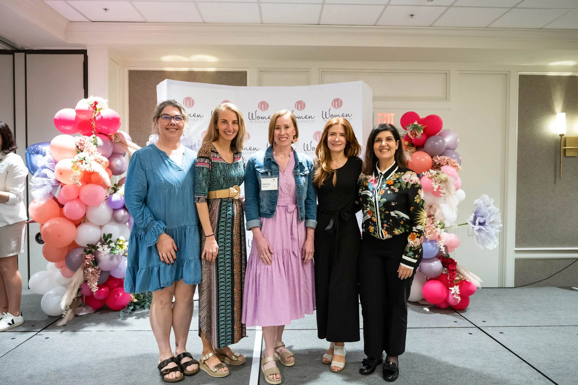 Five women standing together in front of a decorative backdrop with pink, purple, and white balloons and flowers, attending an event called Women in Pediatric Matric. They are smiling and dressed in colorful, casual and semi-formal clothing.