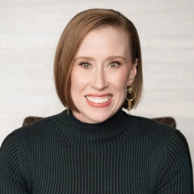 A woman with short, straight, light brown hair, wearing a black ribbed turtleneck sweater, gold hoop earrings, smiling, and sitting in front of a light-colored background.