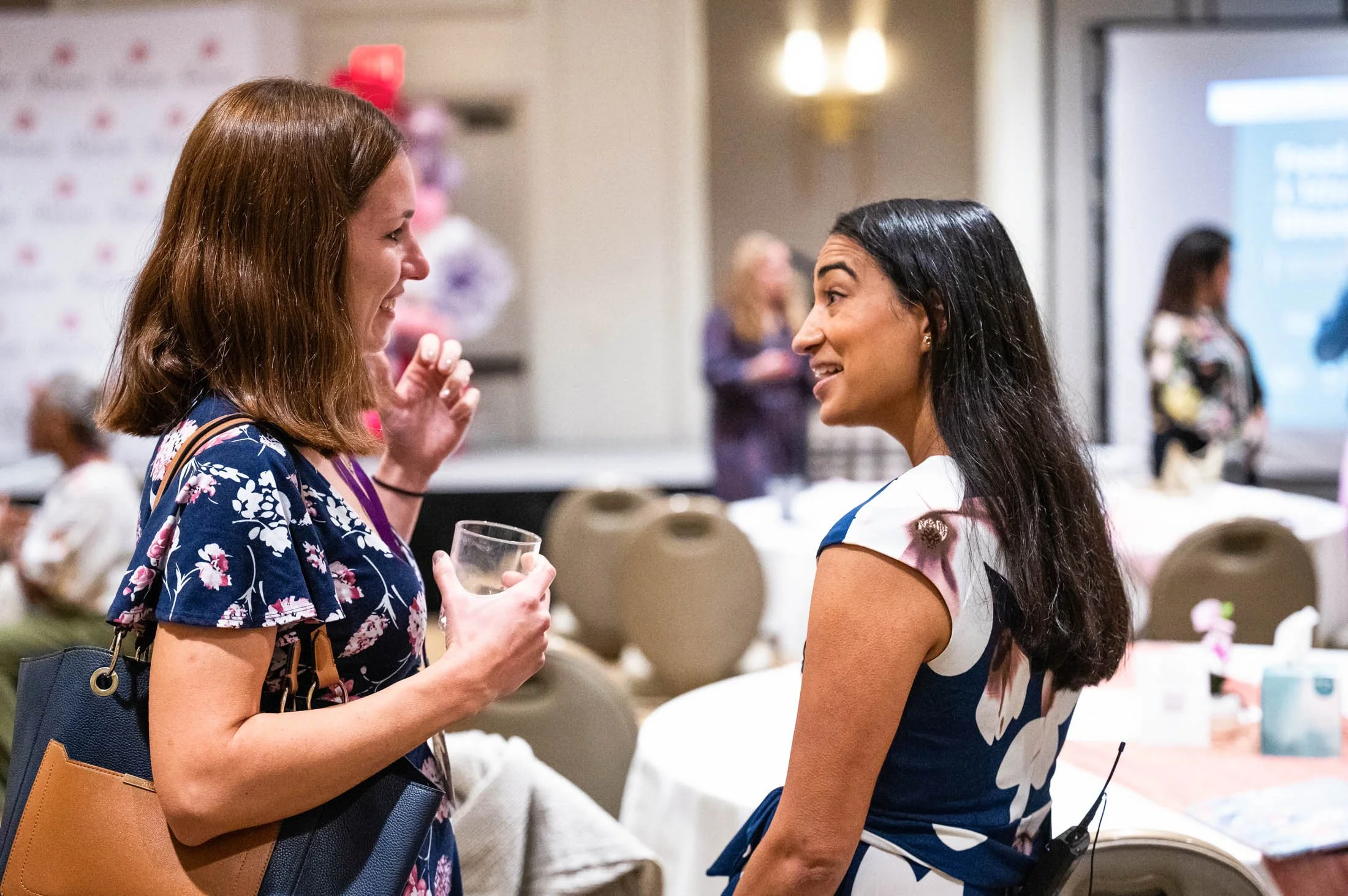 Two women are talking and smiling at each other at an indoor event. One woman has red hair and glasses, and is holding a glass of water. The other woman has long dark hair and is wearing a patterned dress. There are additional people and decorations in the background.