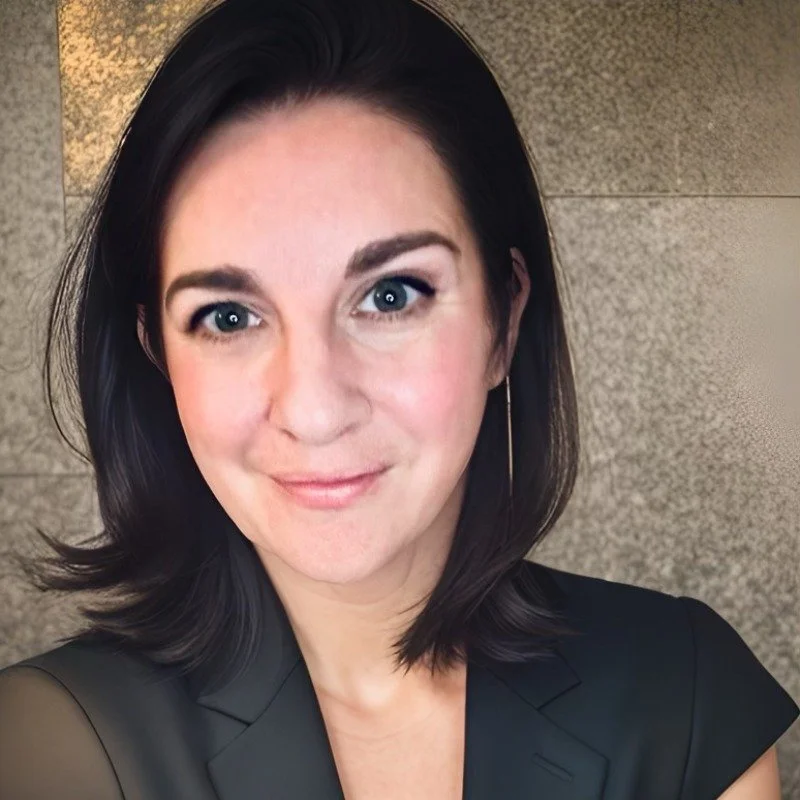 A woman with shoulder-length dark hair, wearing a black blazer, smiling in front of a textured wall.