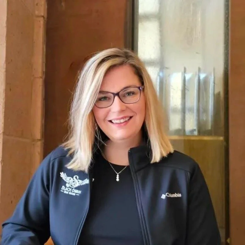 A young woman with blonde hair, glasses, and a black Columbia jacket smiling indoors near a window.