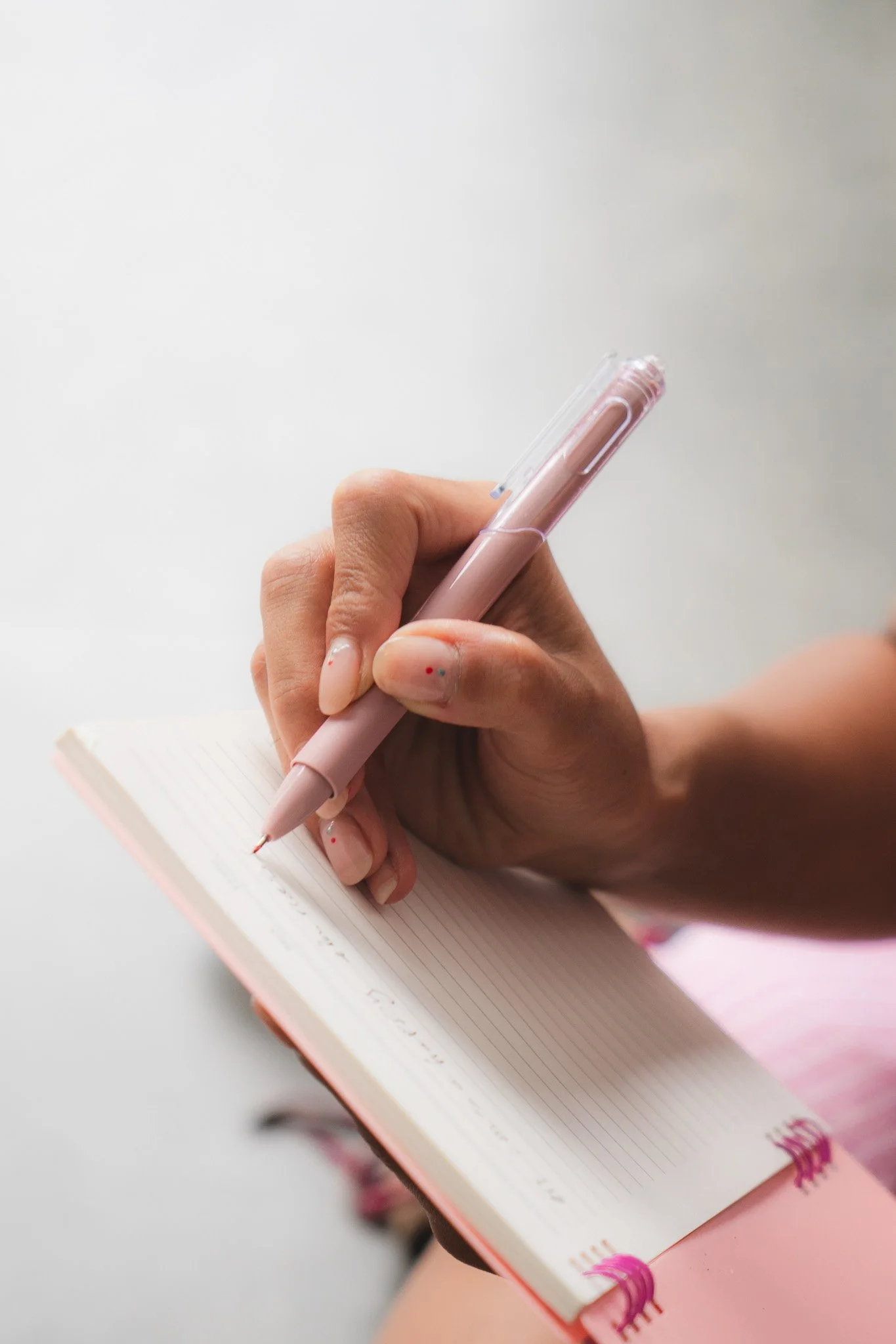 Person holding a pink pen and writing in a small lined notebook with a pink cover, sitting at a white surface.