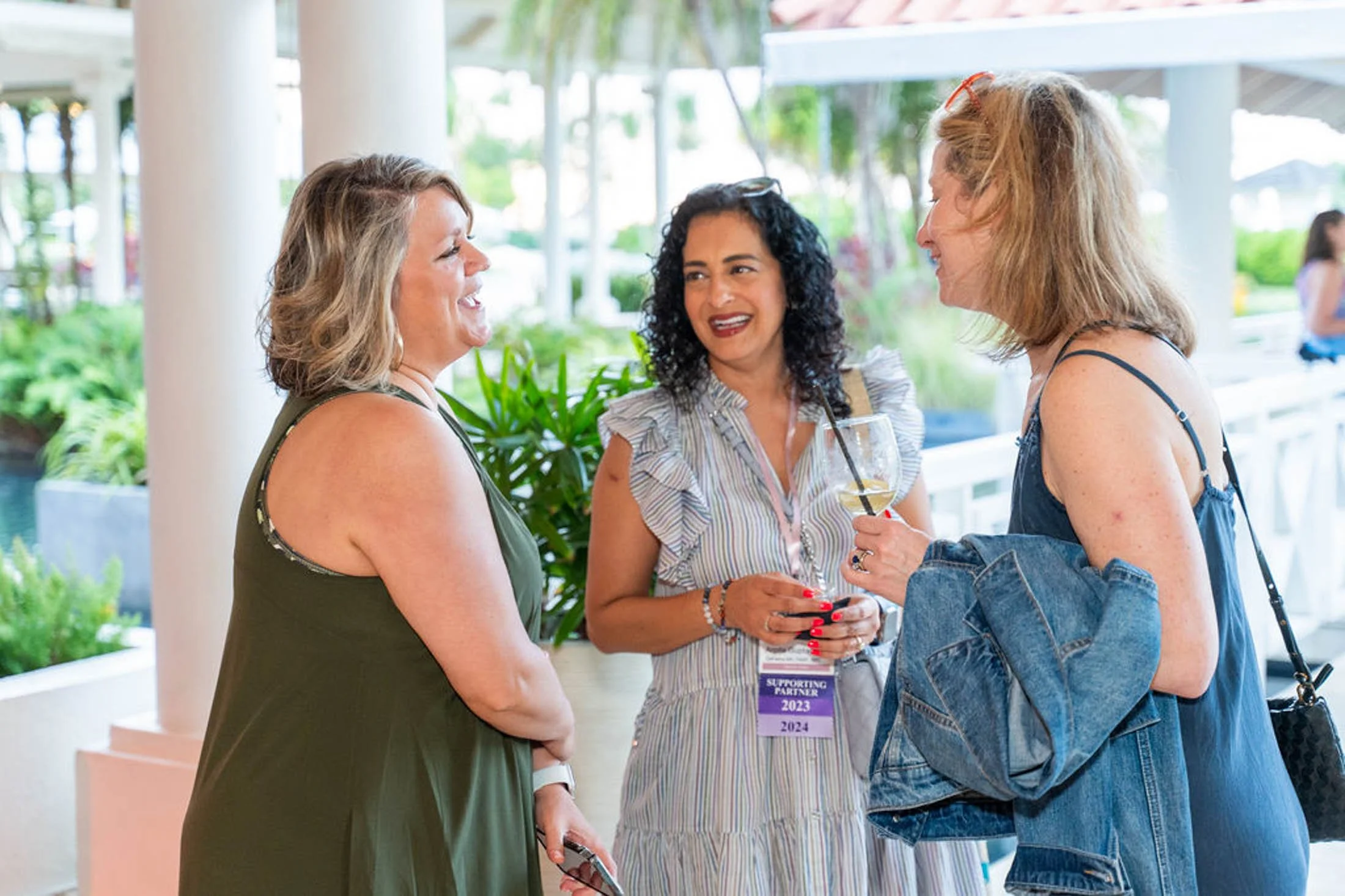 Three women are talking and smiling at each other at an outdoor event. One woman is holding a drink, and they appear engaged in conversation.