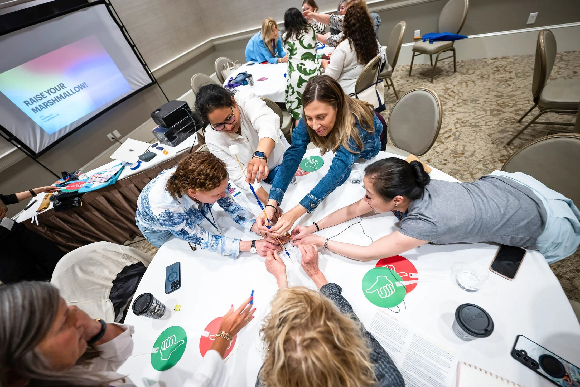 Group of people participating in a team-building activity around a table, connecting wires or cords. Some are reaching across the table, others are watching. A large television screen displays the text 'RAISE YOUR MARSHMALLOW!'. The scene takes place in an indoor conference room with chairs and tables.