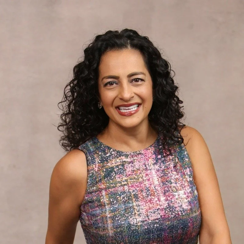 A woman with curly black hair wearing a colorful, patterned sleeveless top and smiling at the camera.