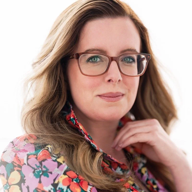 A woman with long, wavy brown hair and glasses with a reddish-brown frame, wearing a floral blouse with red, pink, green, and yellow colors, posing against a white background.