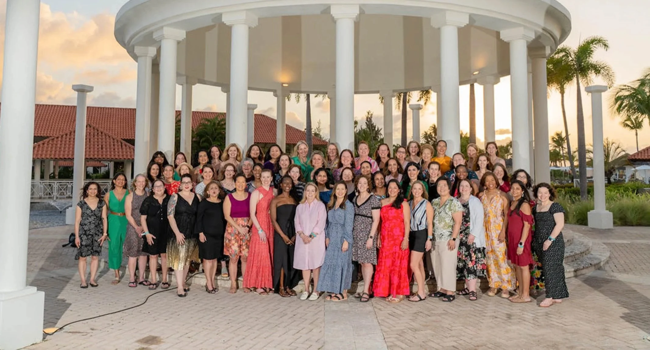 A large group of women standing together outdoors under a white gazebo with columns, dressed in colorful casual and semi-formal clothing, during sunset with palm trees and red-tiled roofs in the background.