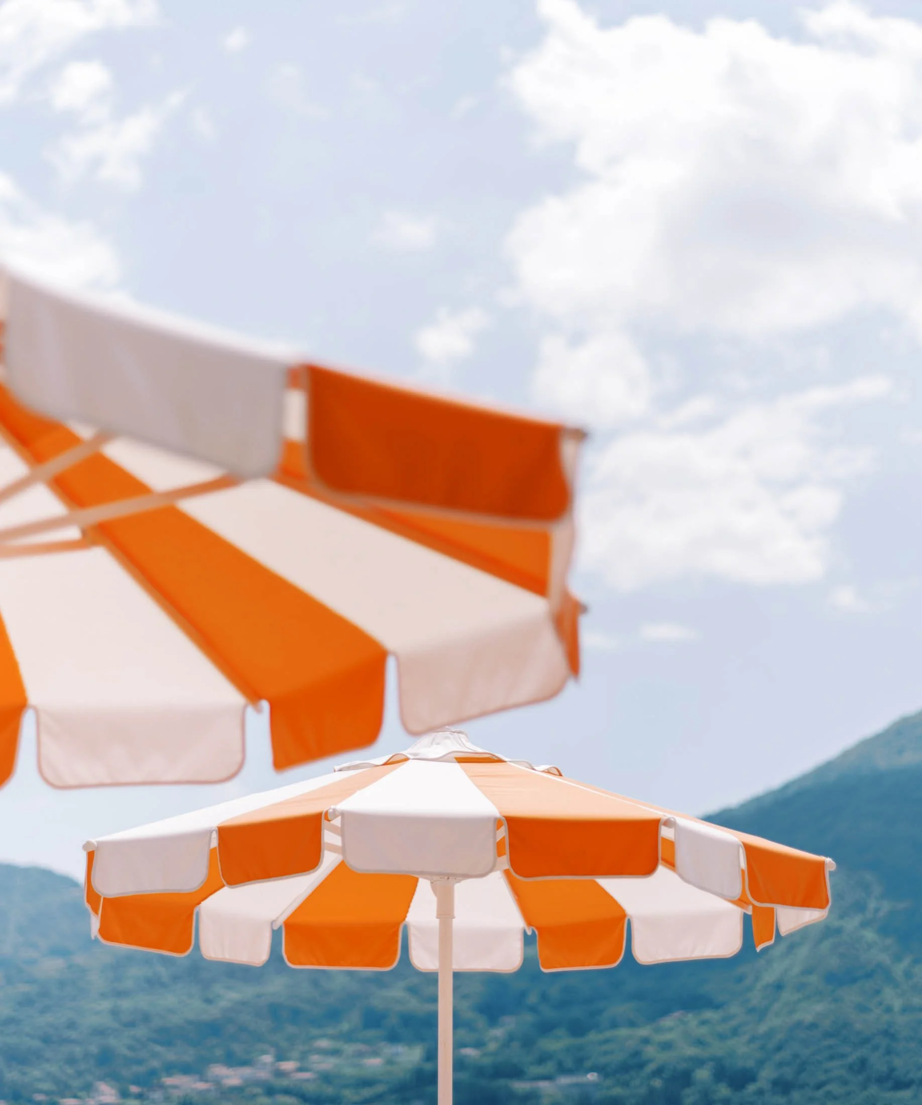 Multiple orange and white striped umbrellas against a blue sky with some clouds and mountains in the background.