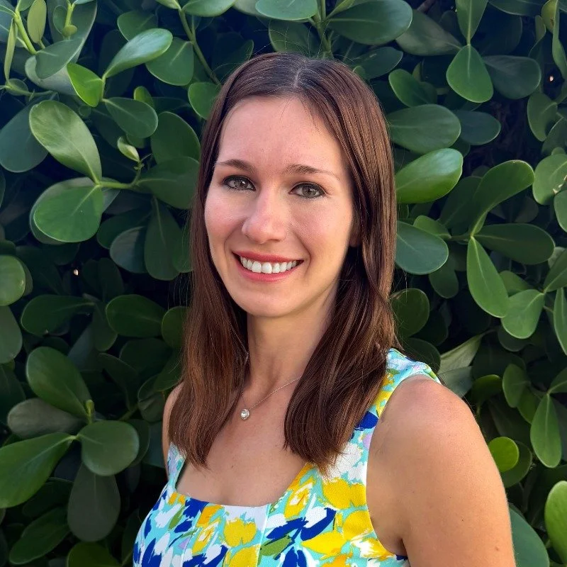 A woman outdoors smiling, wearing a sleeveless floral dress with yellow, blue, and white colors, standing in front of green leafy plants.