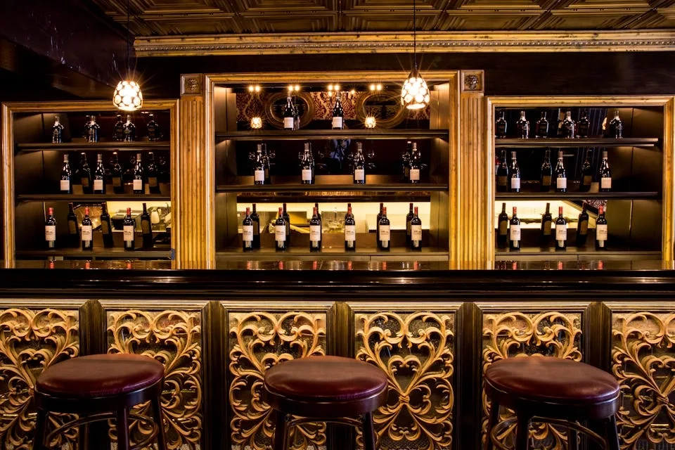A bar with a dark wooden counter and ornate gold detailing, featuring a shelf with wine bottles, decorative ornaments, and hanging pendant lights.
