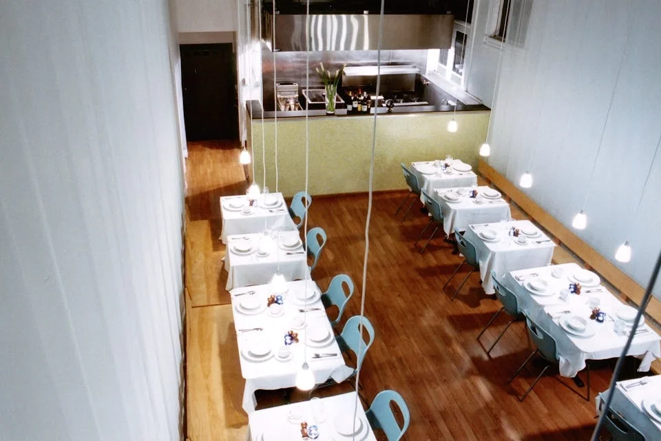 Empty restaurant with white tablecloth-covered tables set with plates, cups, and small decorative items, illuminated by hanging pendant lights, with a view into the kitchen area at the back.