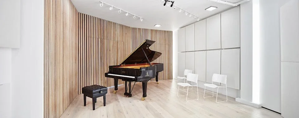 A black grand piano with a matching black bench in a bright, modern music room with wooden wall panels and white chairs.