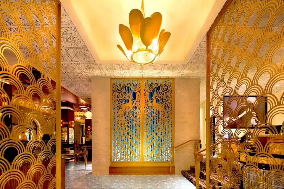 Interior view of a luxury hotel lobby in gold and cream tones with decorative gold metal panels featuring peacock and wave patterns, a lakeside themed wall with a large mural of peacocks, and a gold chandelier hanging from the ceiling.
