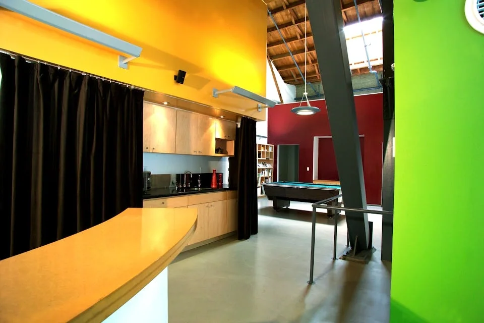 Modern interior with a yellow ceiling, black curtains, wooden cabinets, a bar countertop with a curved edge, a pool table, a red wall, and a green wall, featuring an open ceiling with exposed wooden beams.