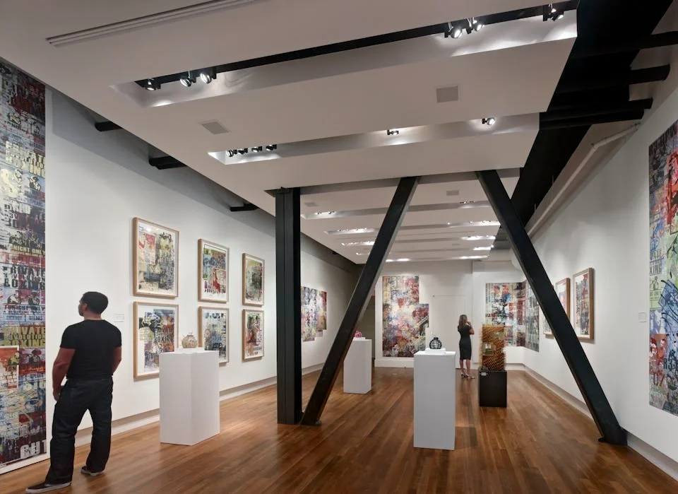 Interior of an art gallery displaying colorful abstract paintings on white walls, with two visitors observing the artwork.