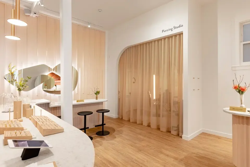 Inside a modern piercing studio with minimalist decor, light wood accents, soft beige curtains, and a white reception counter with jewelry displays and tablets.