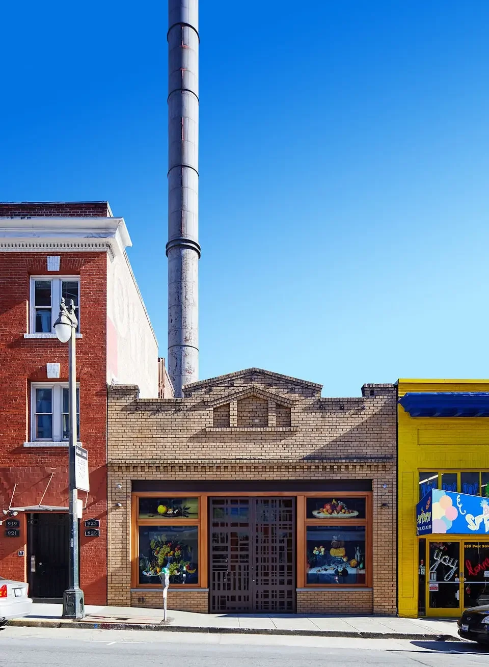 Urban street with brick buildings, storefronts, and a tall industrial chimney against a bright blue sky.