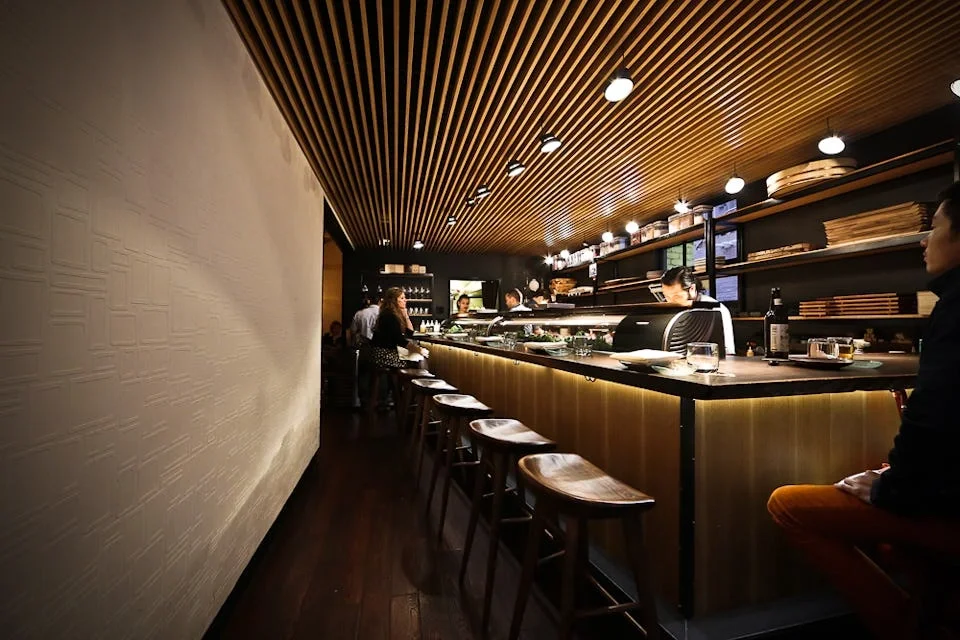 Interior of a modern sushi restaurant with bar seating, chefs preparing food behind the counter, and customers dining.