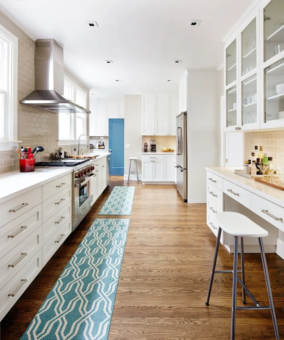 Bright modern kitchen with white cabinets, stainless steel appliances, wooden floors, and patterned blue rugs.