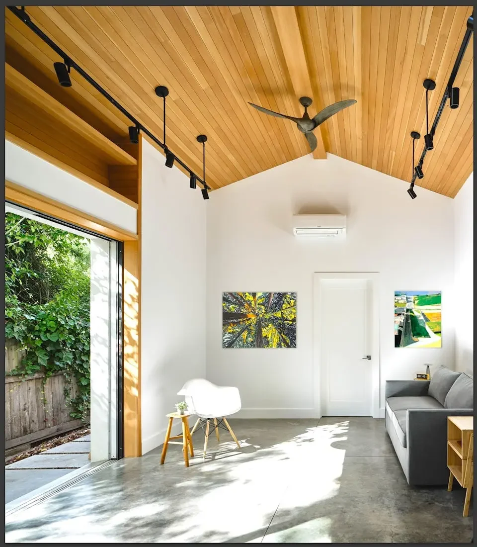 Living room with high wooden ceiling, white walls, large sliding glass door, modern white chair, gray sofa, artwork on walls, ceiling fan, track lighting, and an air conditioning unit.