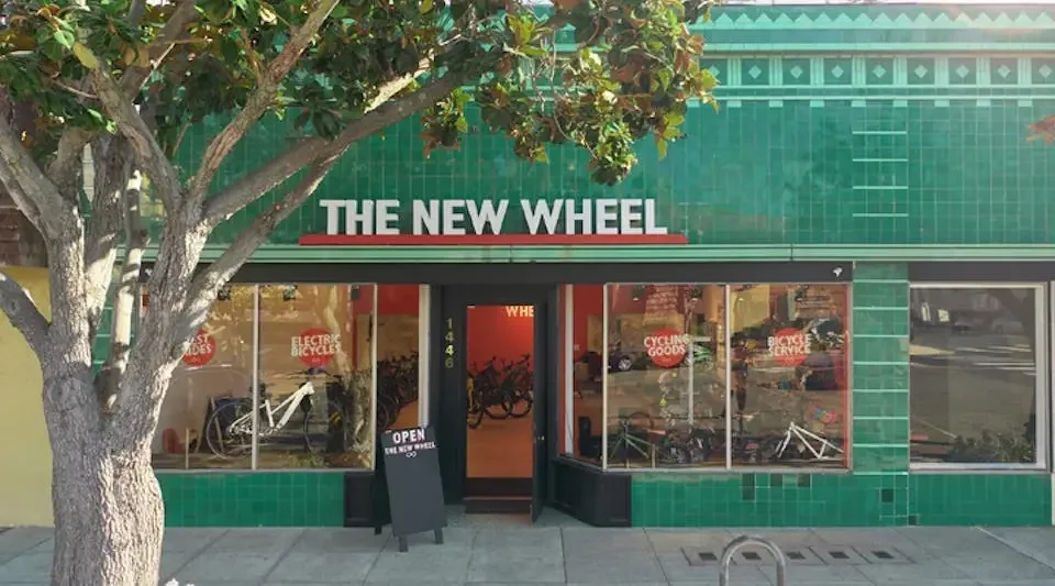 Green storefront with sign 'The New Wheel', large windows showing bicycles inside, and a sidewalk outside with a tree.