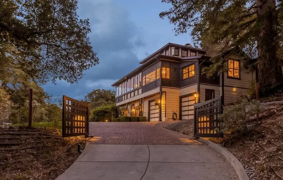 A two-story house on a hill with lights on, surrounded by trees and a decorative gate at the entrance, during dusk.
