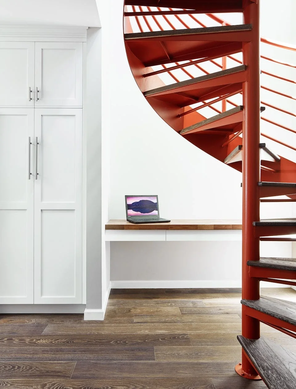 Interior view of a room with white cabinetry, a small desk with a laptop, and a red spiral staircase with wooden steps.