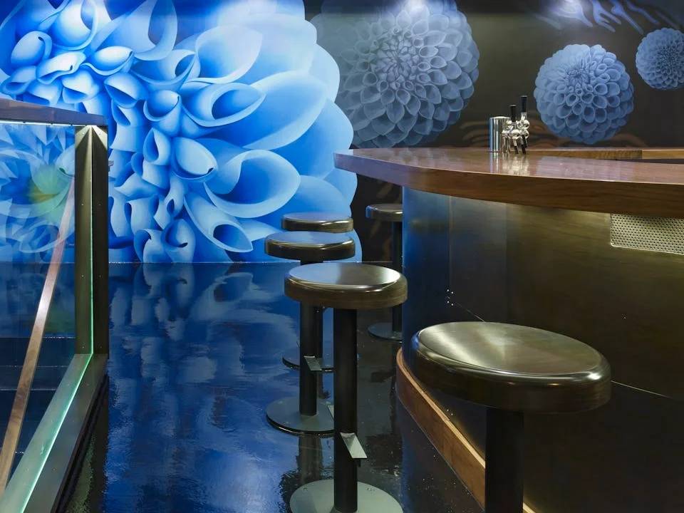 Interior of a modern bar with a curved wooden counter and black bar stools, featuring a large blue floral mural on the wall.