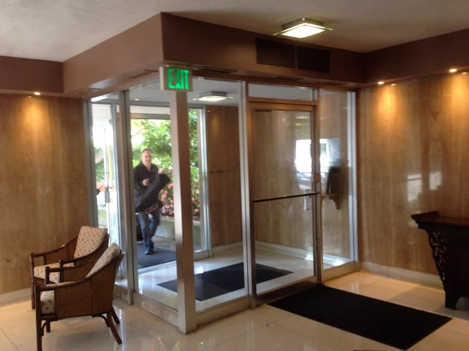 Indoor hotel lobby entrance with glass doors, a person entering, two chairs on the left, and a black rug on the floor.