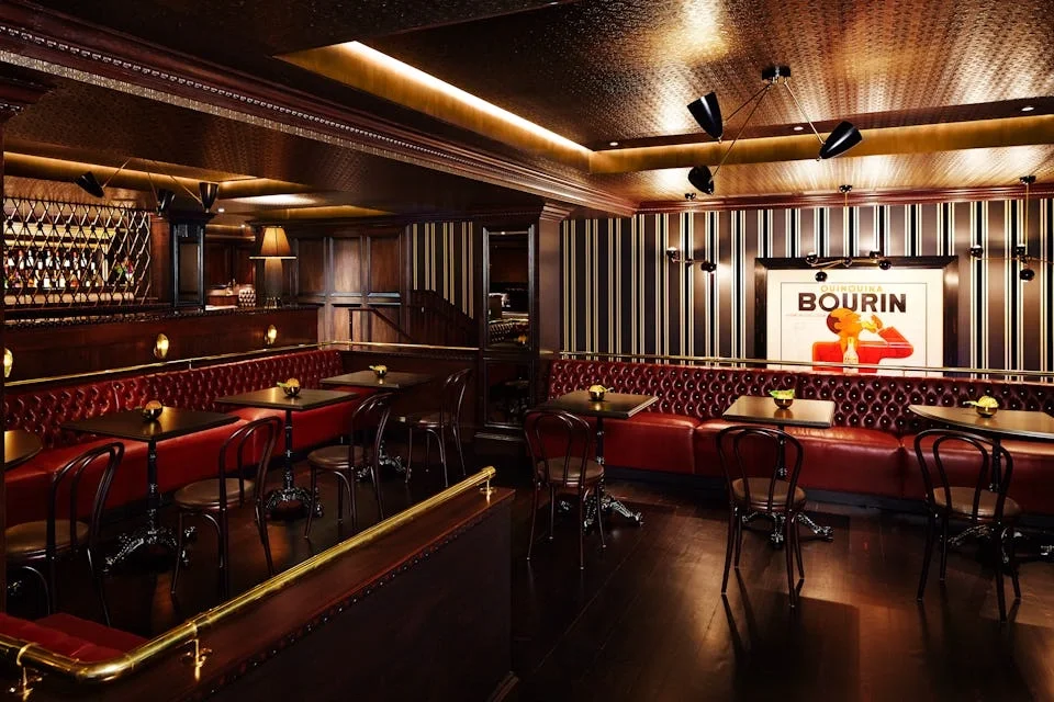 A dimly lit restaurant interior with dark wood furniture, red upholstered seating, and black chairs. There are small table lamps, striped wallpaper on the walls, and a framed vintage cigarette ad poster.