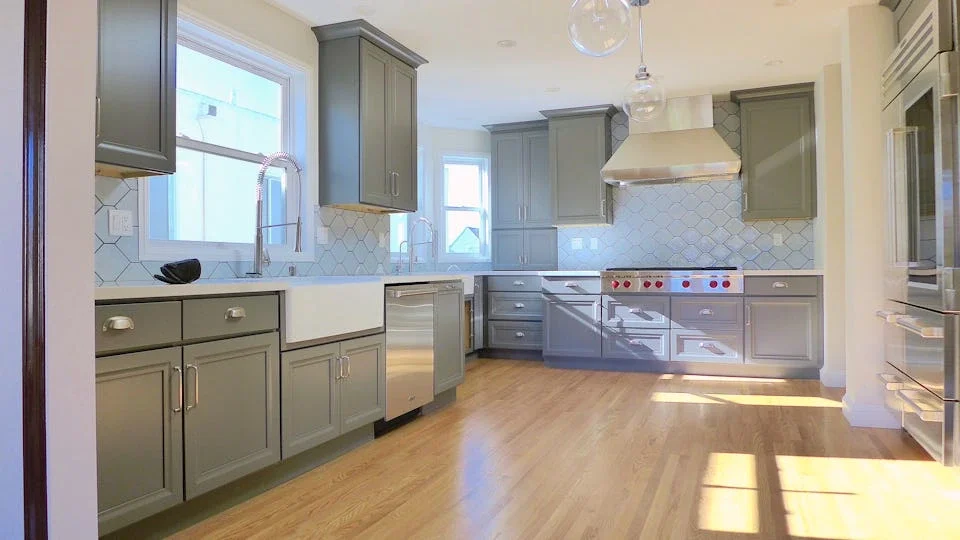 Modern kitchen with green cabinetry, white tile backsplash, large farmhouse sink, wooden floor, and stainless steel appliances.