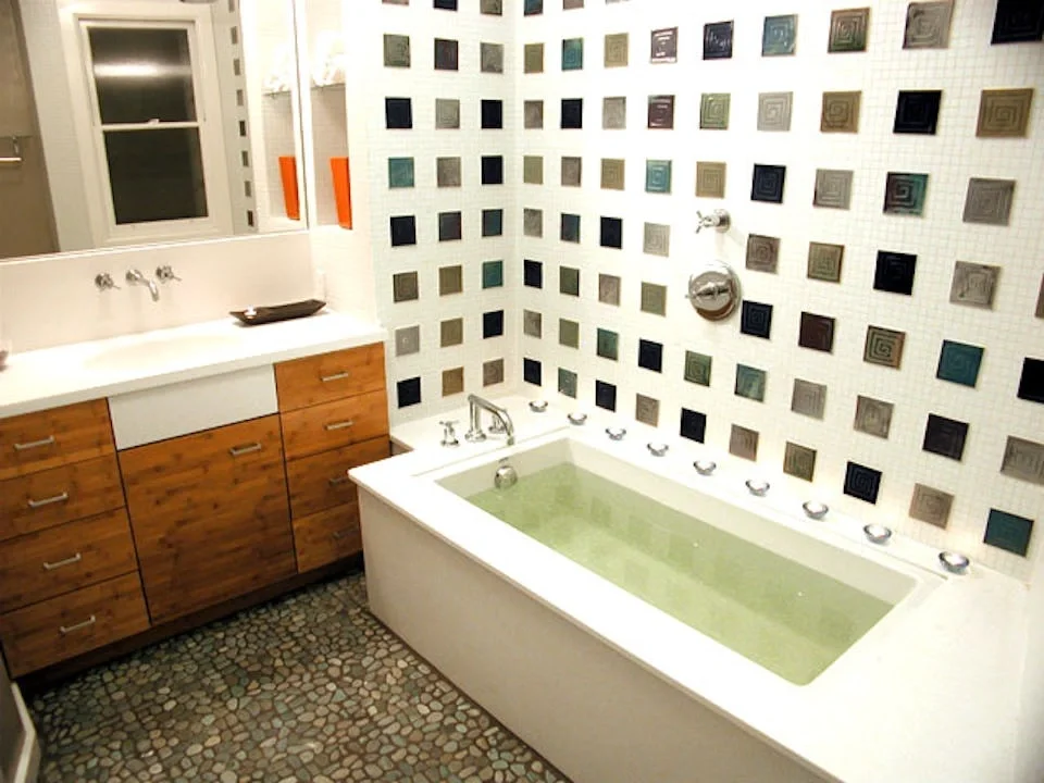 Bathroom with a bathtub filled with green water, a wooden vanity with a white sink and a mirror, a wall with multicolored square tiles, and pebble flooring.