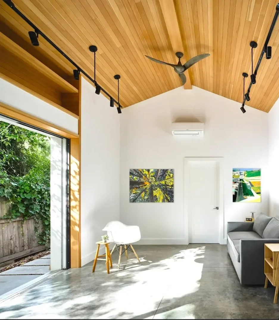 Bright living room with a wooden ceiling, track lighting, a ceiling fan, white walls, an air conditioning unit, artwork on the walls, a gray sofa, a white chair with wooden legs, and a sliding glass door leading outside to greenery.