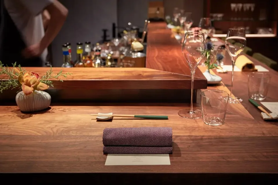A cozy restaurant bar counter with a small flower arrangement, a rolled cloth napkin, glasses, and a menu. In the background, there are bottles of liquor and bar tools.