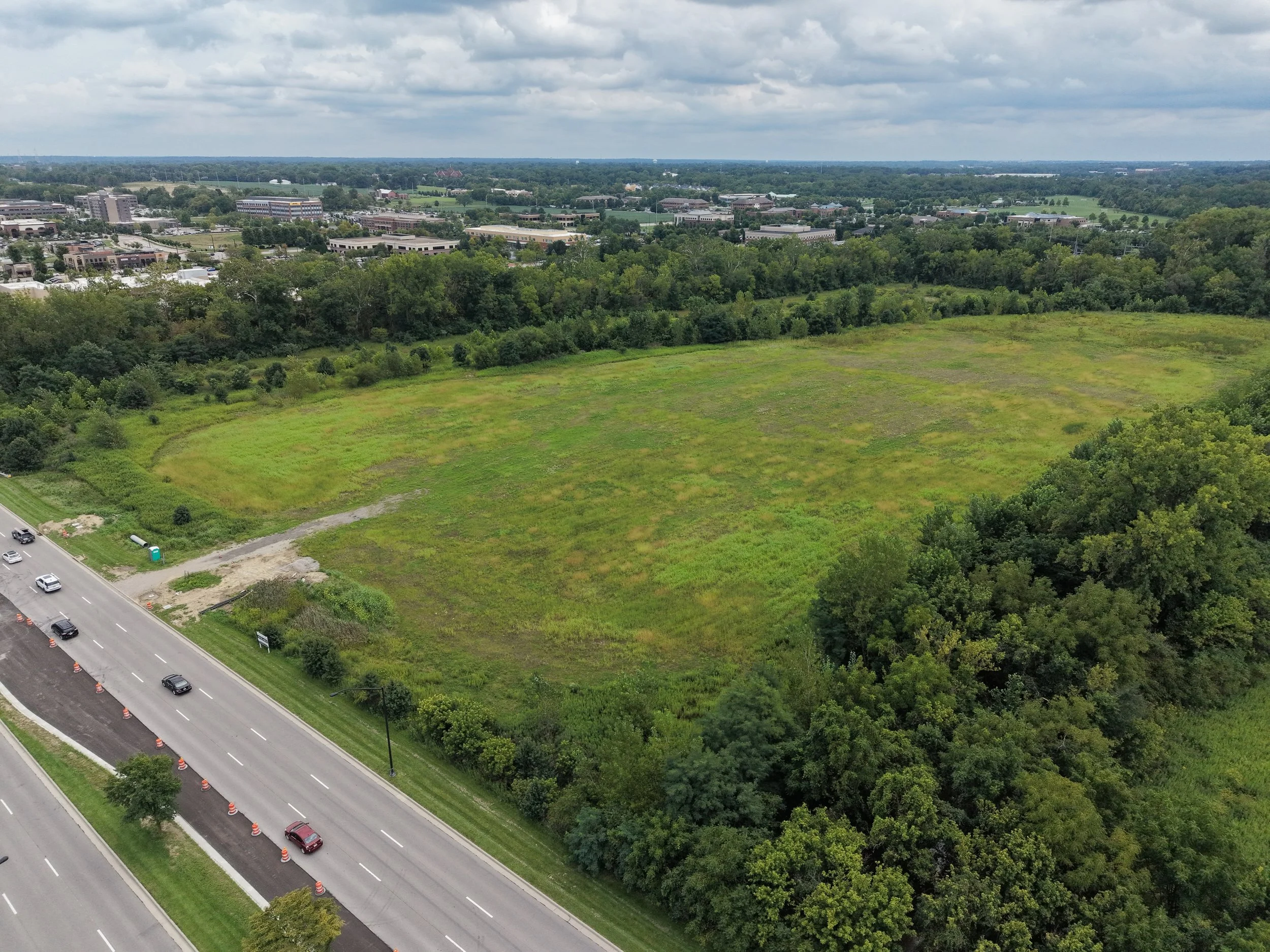 Aerial view of a city landscape with a mix of green spaces, trees, and buildings under a cloudy sky, with a highway and some cars in the foreground.