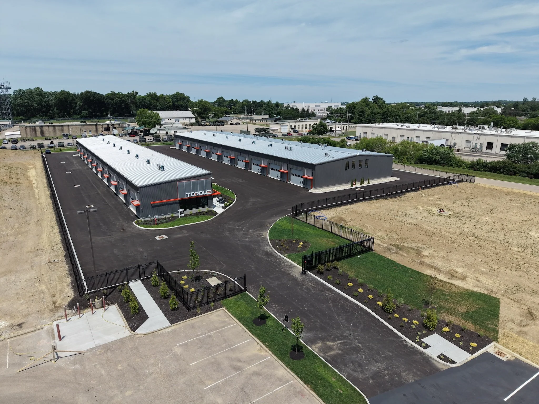 A drone photo aerial view of an industrial or commercial complex with gray buildings, parking lots, and a new landscaped area with small trees and shrubs, under a partly cloudy sky.