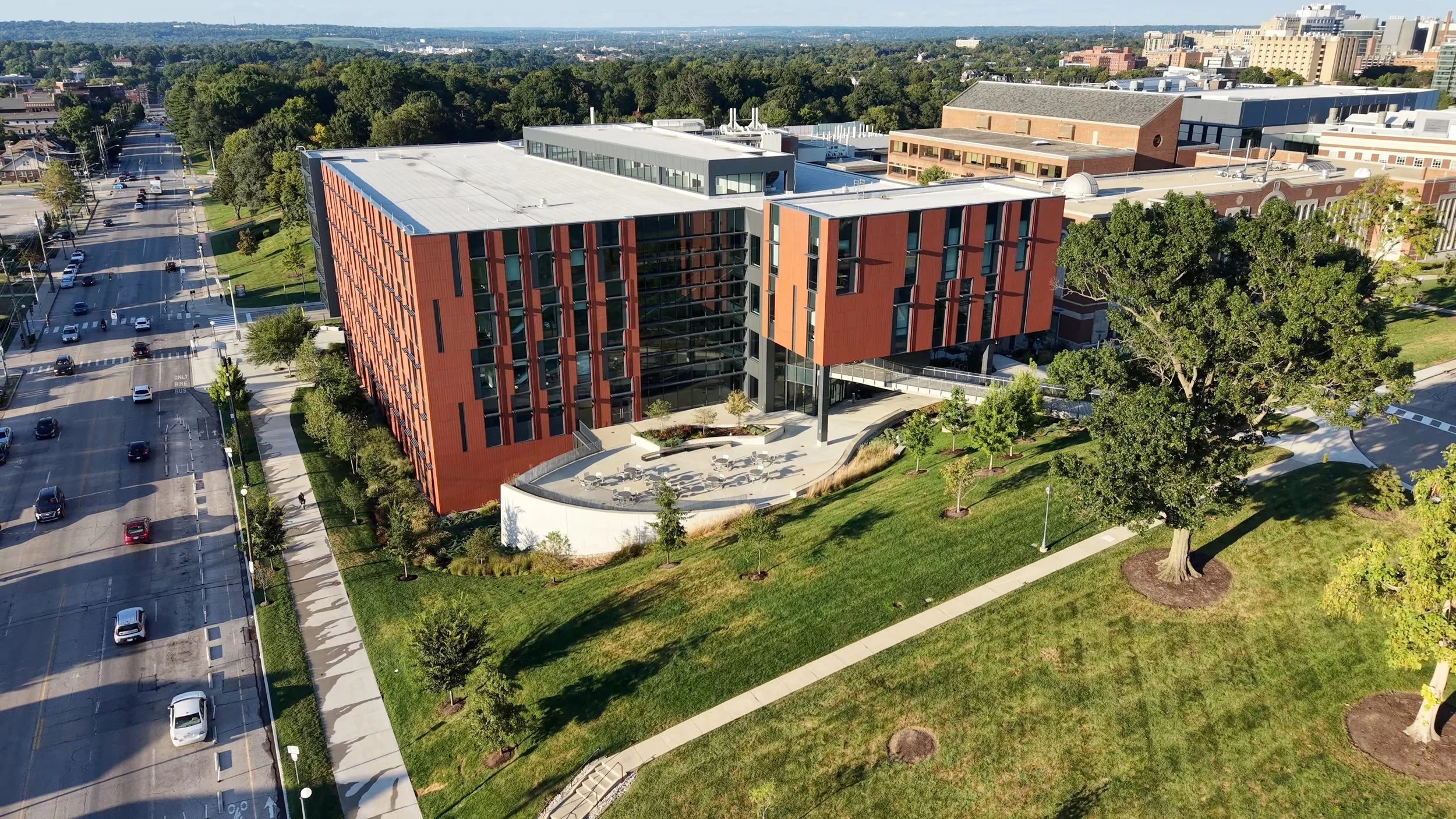 Aerial drone photo view of a modern building with red and black exterior, surrounded by green trees, sidewalks, and roads with moving cars, in an urban area with additional buildings and a distant cityscape.