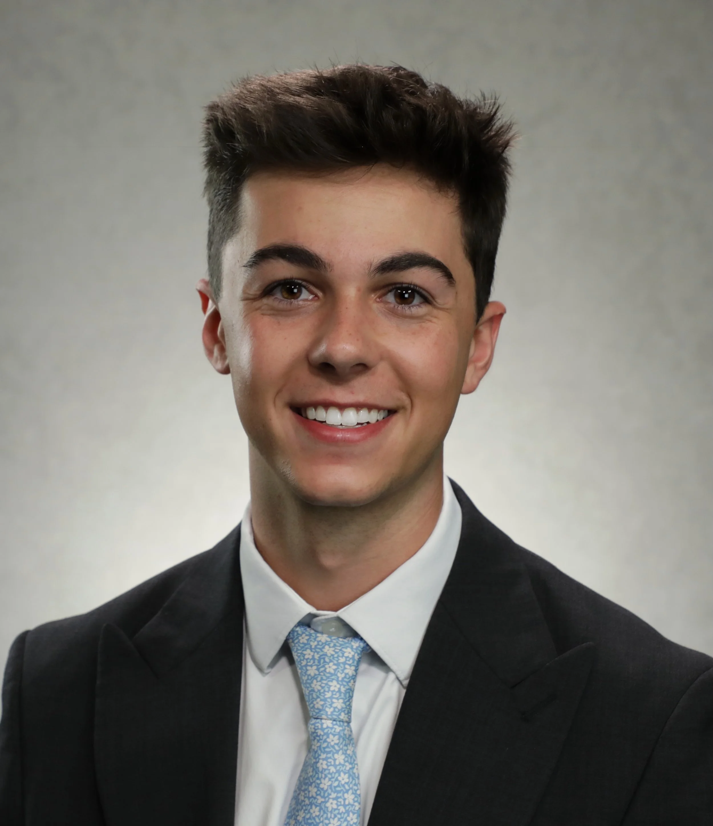 Benjamin Lones is smiling, wearing a dark suit, white shirt, and a light blue patterned tie, with a gray background.