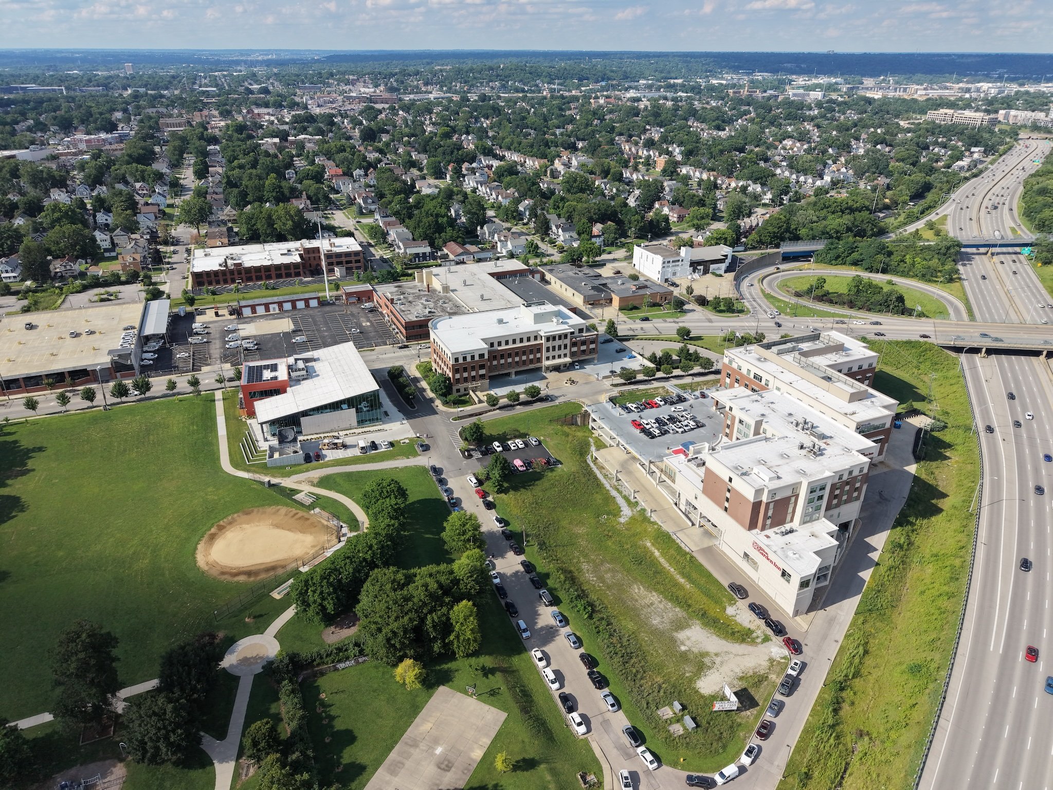 Aerial view of a cityscape including a park with a baseball field, residential neighborhoods with houses and trees, commercial buildings, and a highway with moving cars.