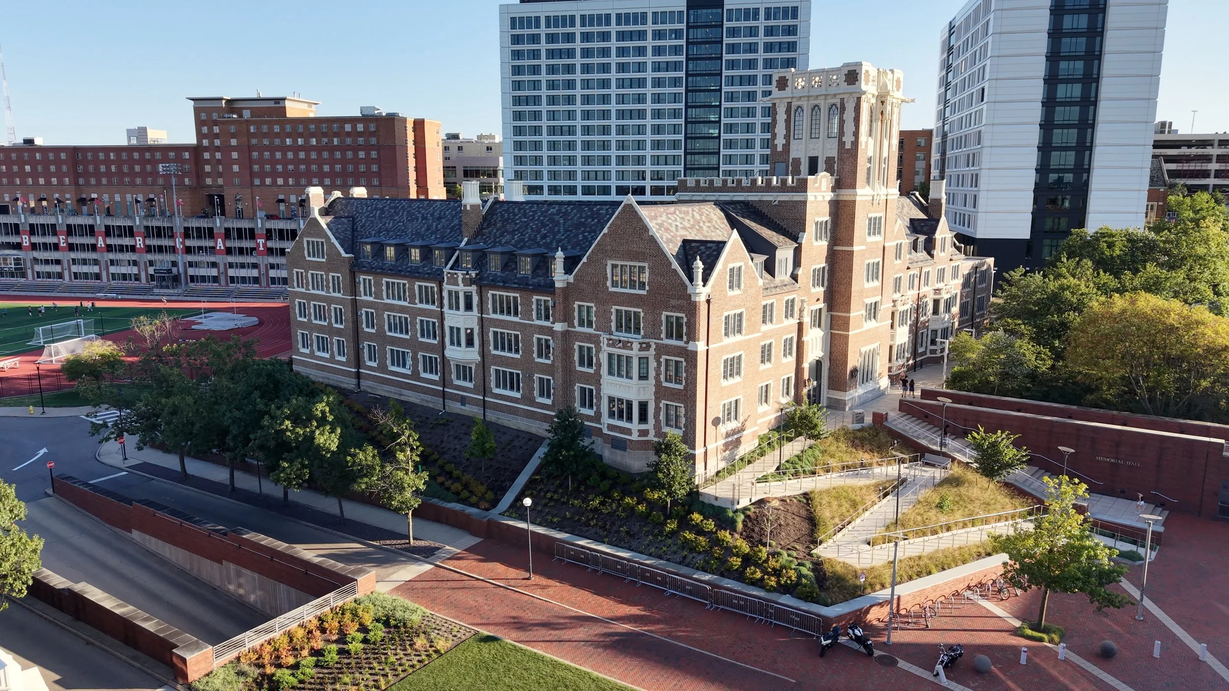 A cityscape with a mix of modern and historic buildings, including a large brick building with white windows, a high-rise with a glass facade, and a stadium with the word 'BEARCATS' on it. There are trees, a walking path, and a few people in the area