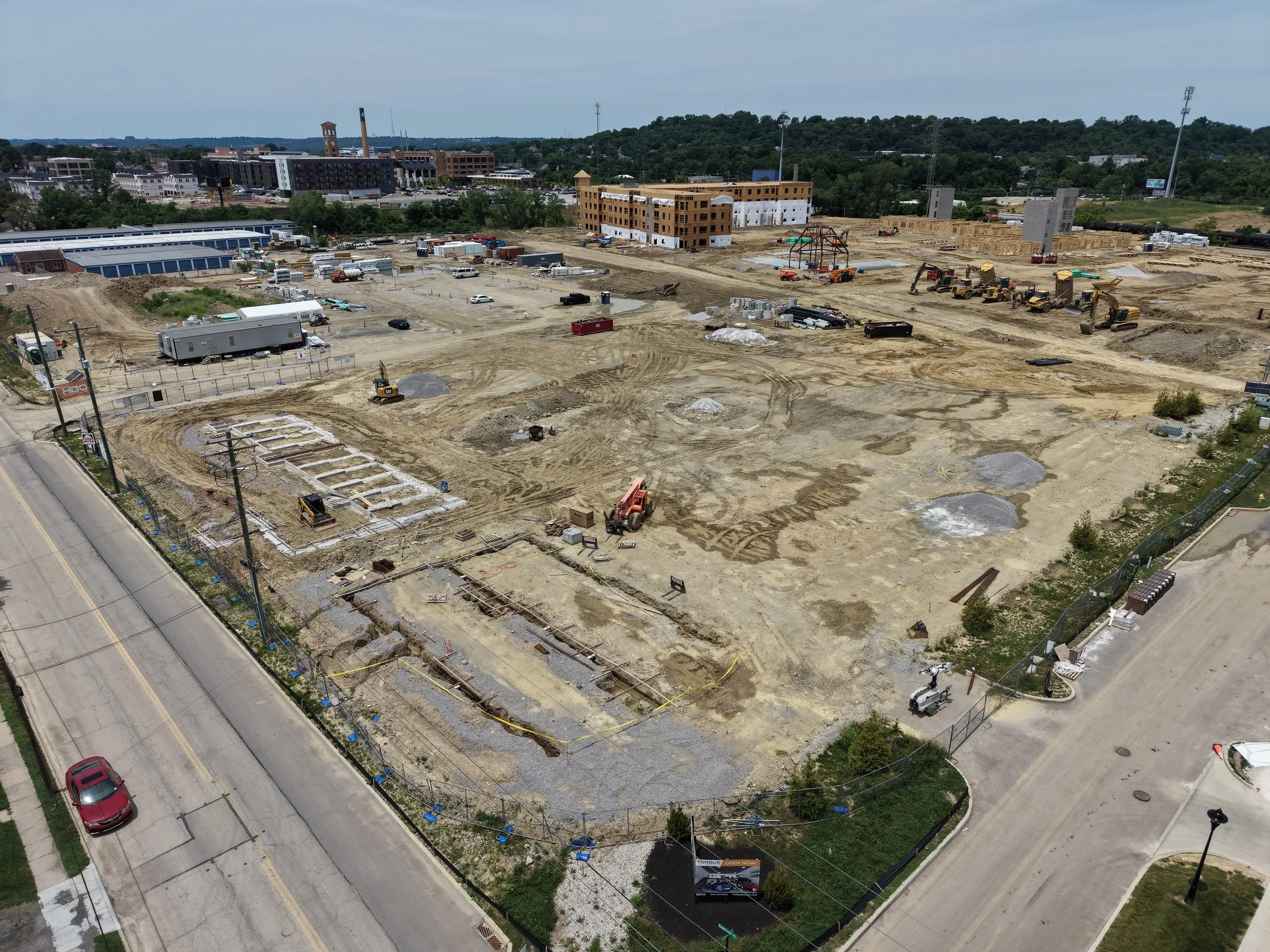 Construction site with multiple equipment and partially built structures, surrounded by a road and nearby buildings in the background.
