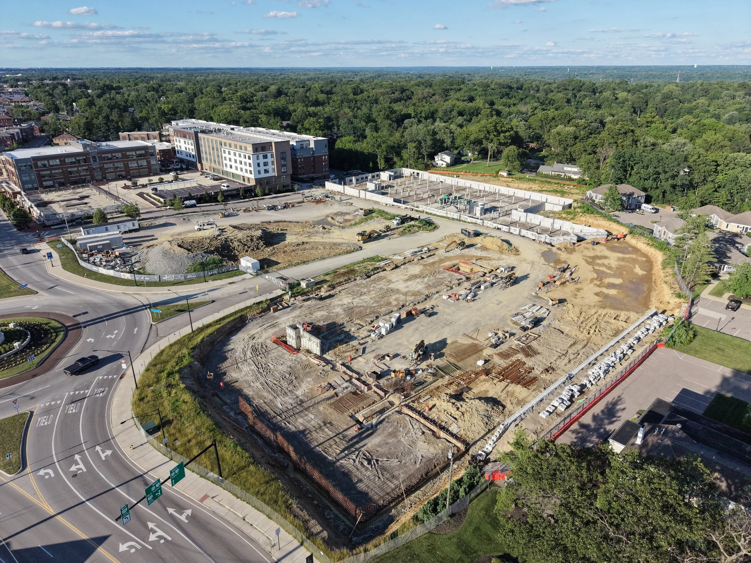 An aerial view of a construction site next to a residential neighborhood, with a wooded area in the background. The site has ongoing excavation, concrete foundations, and construction equipment. Surrounding roads have traffic signs and lanes.