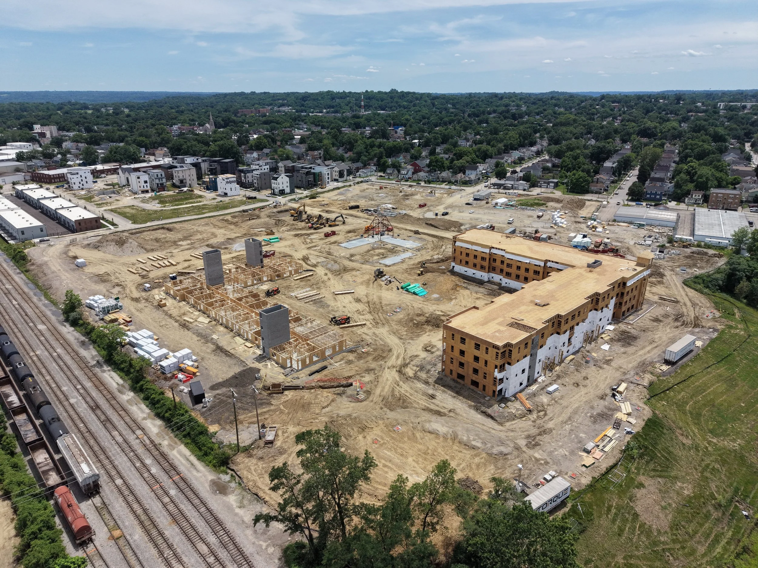 Aerial view of a construction site with buildings under development, surrounded by a neighborhood with houses and trees, and a railway with train cars on the left side.