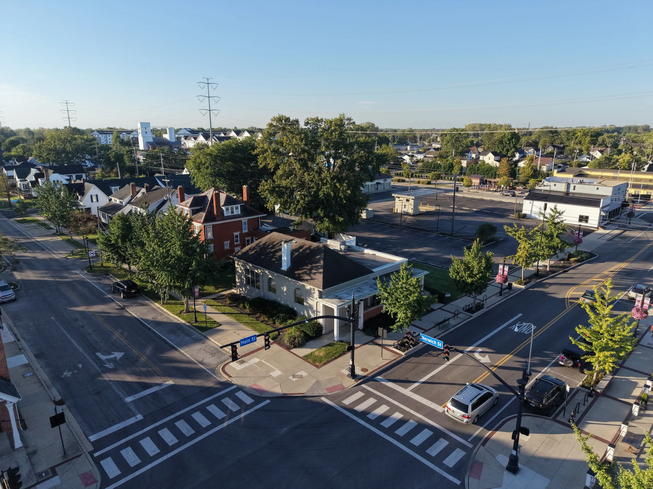 Aerial view of a small town with residential and commercial buildings, streets, and trees, showing a clear sky and bright daylight.