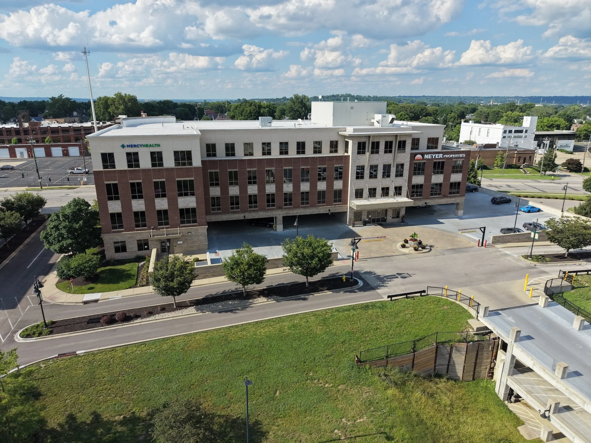 An aerial view of a multi-story building with signage for Mercy Health and Neyer Properties, surrounded by parking lots, trees, and a green lawn, with a partly cloudy sky overhead.